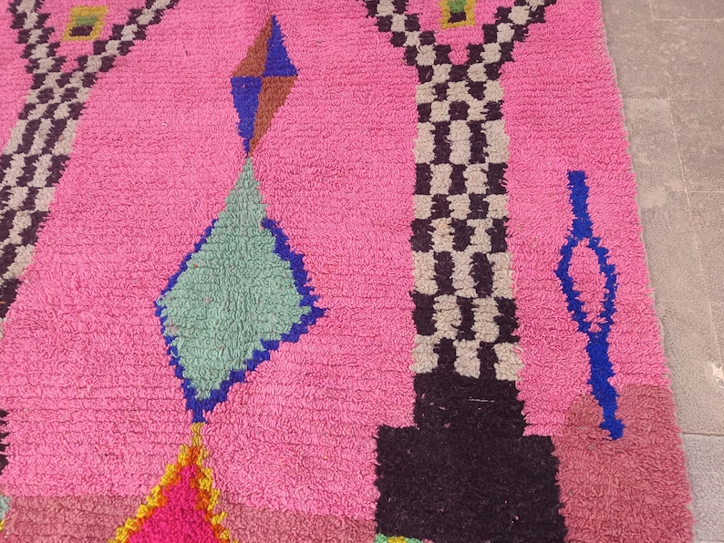 May include: A vibrant pink rug with geometric patterns. The design includes diamond and rectangular shapes in blue, green, brown, and black. The rug has a textured surface and a colorful, eye-catching design.