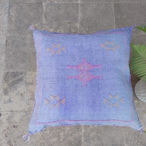 May include: A square, light blue woven pillow with a geometric design in pink and tan. The pillow has a textured appearance and is adorned with tassels at each corner. The pillow is placed on a gray stone surface.