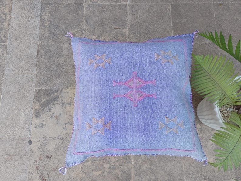 May include: A square, light blue woven pillow with a geometric design in pink and tan. The pillow has a textured appearance and is adorned with tassels at each corner. The pillow is placed on a gray stone surface.