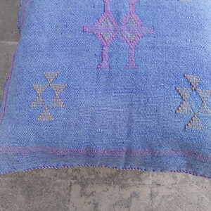 May include: A square, light blue cushion with geometric patterns in pink and beige. The cushion has a textured appearance and a pink border. The design features symmetrical shapes, creating a balanced and decorative look.