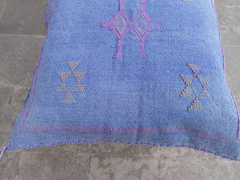 May include: A square, light blue cushion with geometric patterns in pink and beige. The cushion has a textured appearance and a pink border. The design features symmetrical shapes, creating a balanced and decorative look.
