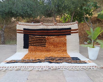 May include: A rectangular rug with a geometric design. The rug features a cream-colored background with black, brown, and orange geometric shapes. The rug has a fringe along the bottom edge. The rug is displayed outdoors.