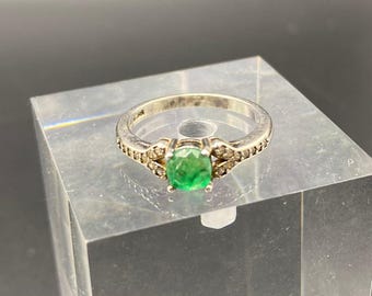 May include: A silver ring featuring a vibrant green emerald gemstone as the centerpiece. The band is adorned with small, sparkling accent stones. The ring is displayed on a clear, angled surface, showcasing its design.