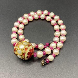 May include: A double-strand necklace featuring creamy white pearls and small, vibrant red beads. The necklace includes a large, ornate pendant with gold accents, white and red enamel, and a decorative clasp.