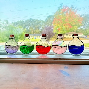 May include: Five stained glass potion bottles with cork stoppers, each filled with a different colored liquid: purple, green, red, pink, and blue. The bottles are displayed on a windowsill with a view of trees and houses in the background.