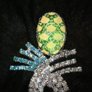 May include: A rhinestone brooch in the shape of a pineapple. The top is oval-shaped with green and yellow rhinestones. The body is silver with clear rhinestones and blue-green accents. The text "ALABAMA WHOLESALE CROWNS LLC" is at the top.