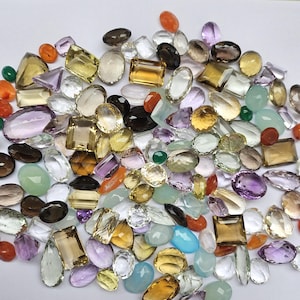 May include: A selection of assorted gemstones in diverse shapes and hues, including yellow, purple, clear, and brown. The gemstones are spread across a white surface, highlighting their facets and clarity. The collection features oval, rectangular, and teardrop cuts.