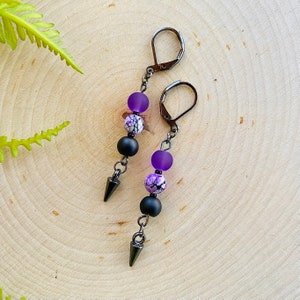May include: A pair of dangle earrings with black metal hooks, purple and white patterned beads, and black metal tips.
