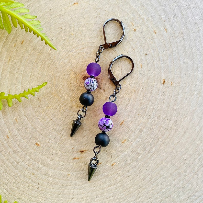 May include: A pair of dangle earrings with black metal hooks, purple and white patterned beads, and black metal tips.