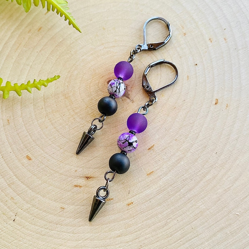 May include: A pair of dangle earrings with black and purple beads. The beads are round and have a marbled pattern. The earrings have a silver-tone finish and a spike at the bottom.