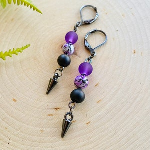May include: A pair of dangle earrings with black and purple beads and a black metal spike at the bottom.