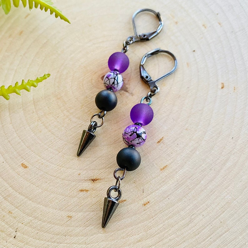May include: A pair of dangle earrings with black and purple beads and a black metal spike at the bottom.
