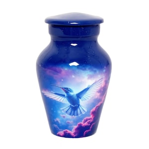 Puede incluir: Urna funeraria azul con acabado brillante. La urna presenta un colibrí en vuelo sobre un fondo de nubes y cielo estrellado. La urna tiene una tapa a juego.