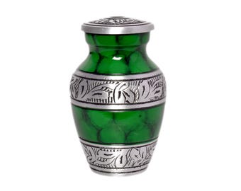 Puede incluir: Una pequeña urna verde con tapa plateada y bandas decorativas. La urna tiene un acabado verde veteado y bandas plateadas con un diseño de hojas. Una pequeña urna decorativa para recuerdos.