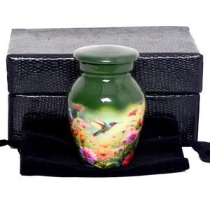 Può includere: Urna in ceramica verde con un design di colibrì e fiori. L'urna è presentata con un sacchetto di velluto nero e una scatola nera strutturata. Un piccolo contenitore decorativo.