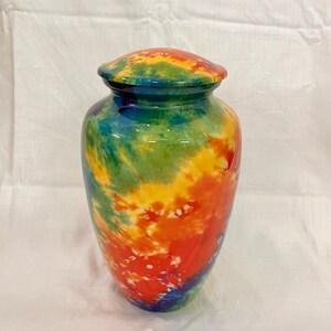 May include: A cylindrical cremation urn featuring a vibrant tie-dye pattern with blended swirls of red, orange, yellow, green, and blue. The vessel has a smooth, glossy finish and includes a matching rounded lid that sits securely on the top opening.