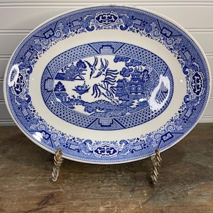 May include: Oval blue and white ceramic platter with a Willow pattern design. The platter features a central scene with a bridge, birds, and pagodas, surrounded by a detailed border. The platter is displayed on a gold-colored metal stand.