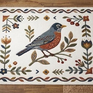 Könnte beinhalten: Rechteckiger weißer Teppich mit detailliertem Stickdesign. Ein blauer, grauer und orangefarbener Vogel sitzt auf einem Ast in der Mitte. Der Rand zeigt Blumen- und Blattmotive in Grün-, Braun- und Blautönen. Der Teppich liegt auf einem Holzboden.