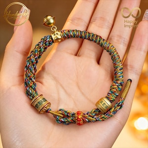Puede incluir: Pulsera trenzada con un dije de campana y detalles dorados. La pulsera presenta un diseño tejido multicolor con hilos rojos, azules, verdes y amarillos. Tiene cuentas cilíndricas doradas con caracteres chinos.
