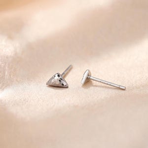 Könnte beinhalten: Ein Paar silberfarbene Ohrstecker in kleiner Dreiecksform mit einer texturierten, facettierten Oberfläche. Jeder Ohrstecker verfügt über einen dünnen Metallstift an der Rückseite, der für das Ohrloch bestimmt ist und glänzt.