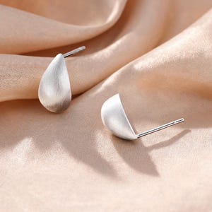 Pode incluir: Um par de brincos de tarracha em tom prateado com um acabamento mate escovado. Cada brinco tem uma forma distinta de gota ou semicírculo com um pino de metal fino que se estende da parte de trás, concebido para fixação no lóbulo.
