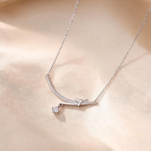 Puede incluir: Un collar de tono plateado con una cadena fina y un colgante de barra curva. La barra está decorada con pequeñas piedras claras y un detalle de nudo central. Una piedra redonda y clara cuelga del nudo, añadiendo un brillo sutil a la pieza.