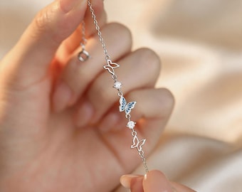 May include: A delicate silver bracelet featuring a series of butterfly charms. The bracelet includes openwork butterfly designs and butterflies with blue enamel wings, interspersed with small, clear gemstones. The bracelet is held against a soft, neutral background.