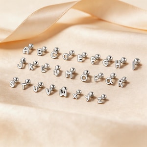 May include: A collection of twenty-six silver-toned metal stud earrings, each featuring a single letter of the alphabet from A to Z. Each earring is designed with a small spherical backing and a flat, uppercase letter front in a clean font.