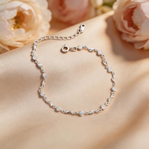 Op de afbeelding: Een delicate zilveren armband met heldere, ronde edelstenen. De armband heeft een herhalend patroon van gebogen schakels en kleine, sprankelende stenen. De sluiting is zichtbaar en de armband ligt op een zacht, perzikkleurig oppervlak.