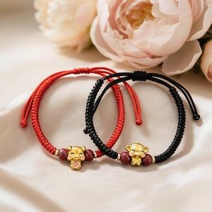 Peut inclure: Deux bracelets tressés, un rouge et un noir, chacun orné d'un charm doré et de perles rouges. Les charms représentent un cochon et un singe. Les bracelets sont présentés sur une surface blanche avec des fleurs roses en arrière-plan.