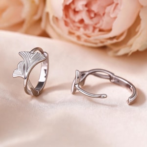 May include: A pair of silver-toned hoop earrings featuring a delicate ginkgo leaf design. The earrings have a polished metallic finish with visible vein detailing on the leaf motifs. One earring is shown upright while the other is lying flat.