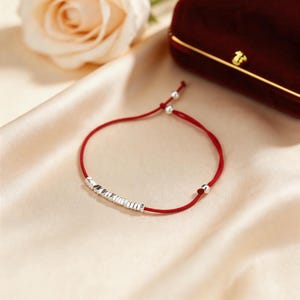 May include: A red string bracelet featuring a row of small, silver-coloured beads. The bracelet has adjustable sliding knots and small silver beads at the ends. The background is a soft, neutral colour.