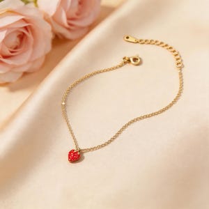 Peut inclure: Un bracelet doré délicat avec un petit charme de fraise rouge. La fraise a des détails de graines blanches. Le bracelet est sur une surface satinée avec des roses roses en arrière-plan.