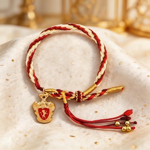 May include: A braided bracelet with red, white, and gold threads. A gold-colored charm with a red crest and a tassel with gold beads hangs from the bracelet. The bracelet is displayed on a white, textured surface.