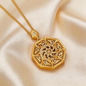 May include: Gold pendant necklace with an elaborate geometric design. The pendant has a central spiral pattern with clear and black accents. The necklace chain is gold, and the background is a soft, cream-coloured fabric.