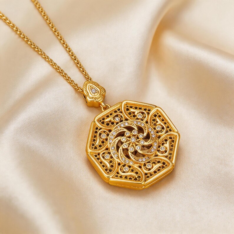 May include: Gold pendant necklace with an elaborate geometric design. The pendant has a central spiral pattern with clear and black accents. The necklace chain is gold, and the background is a soft, cream-coloured fabric.