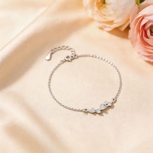 May include: A delicate silver bracelet with a floral design. The bracelet features a series of small, interconnected links and a decorative centerpiece with a cluster of stylized flowers. The bracelet is displayed on a satin surface.