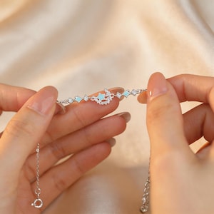 Könnte beinhalten: Ein filigranes Silberarmband mit einem himmlischen Design. Das Armband hat kleine, hellblaue rautenförmige Akzente und einen zentralen Anhänger mit einem Mond- und Sternmotiv, verziert mit klaren Kristallen. Das Armband wird von zwei Händen gehalten.