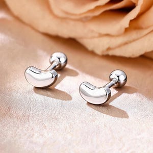 May include: A pair of silver-toned stud earrings featuring a smooth, curved, bean-shaped design. Each earring has a polished metallic finish and a small, round ball backing attached to a straight post, creating a clean and reflective appearance.