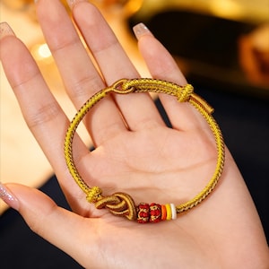 May include: Gold-colored bracelet with intricate knot details and a series of red and yellow beads. The bracelet is displayed on a hand, showcasing its design. The bracelet is a piece of jewelry.