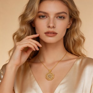 May include: A gold pendant necklace featuring an intricate, geometric design. The octagonal pendant has a detailed, swirling pattern at its centre. The necklace is worn by a person with blonde hair and a cream-coloured top.