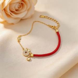 Könnte beinhalten: Goldfarbenes Armband mit rotem Kordelabschnitt. Das Armband hat einen kreisförmigen Anhänger mit einem rautenförmigen Muster und eine kleine, baumelnde Glocke. Ein kleiner, rautenförmiger Stein ist ebenfalls auf der Kette vorhanden.