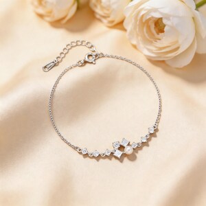 Puede incluir: Una delicada pulsera de plata con una serie de pequeñas piedras cuadradas transparentes y una sola perla. La pulsera tiene una cadena ajustable y un cierre seguro. El fondo es de color neutro.