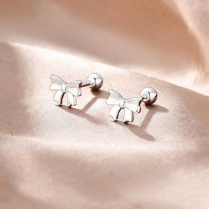 May include: A pair of silver-toned stud earrings featuring a bow design. Each earring consists of a polished bow-shaped front piece connected to a straight post with a small spherical ball backing, creating a cohesive and reflective metallic appearance.