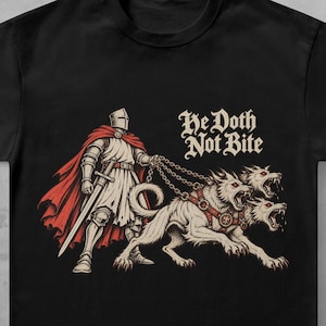 Może przedstawiać: Czarny t-shirt z krótkim rękawem z nadrukiem przedstawiającym rycerza w zbroi z czerwonym płaszczem, trzymającego łańcuchy przyczepione do trzygłowego psa. Napis "He Doth Not Bite" wykonano białą czcionką w stylu gotyckim nad ilustracją psa.