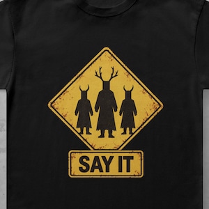 May include: A black short-sleeved t-shirt featuring a yellow diamond-shaped road sign graphic with a distressed texture. The graphic depicts three silhouetted figures, the centre one with antlers, above a rectangular sign reading SAY IT.
