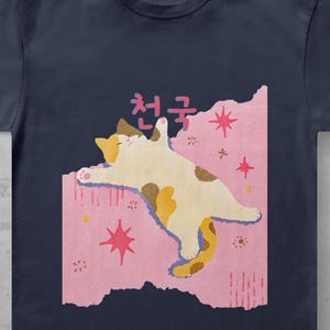 May include: A navy blue short-sleeve t-shirt featuring a graphic of a calico cat lying on a pink rectangular background. The cat is depicted with brown and white patches, and the design includes pink star shapes and the Korean text 천국.