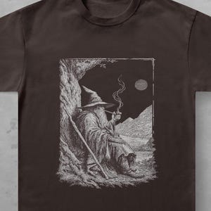 Op de afbeelding: Een donkerbruin t-shirt met korte mouwen en een gedetailleerde witte lijntekening van een bebaarde tovenaar met een puntmuts die onder een boom zit, een pijp rookt, met een staf ernaast en een maan zichtbaar in de nachtelijke hemel.