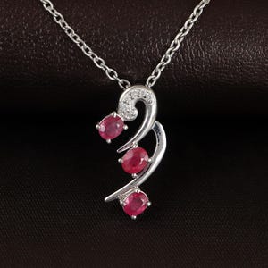 Op de afbeelding: Een zilveren hangerketting met drie ovale, levendige rode edelstenen. De hanger heeft een vloeiend, abstract ontwerp met een kleine cluster van heldere stenen aan de bovenkant. De ketting is van delicaat zilver.