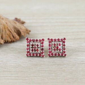 Peut inclure: Une paire de boucles d'oreilles carrées avec un sertissage argenté. Chaque boucle d'oreille est ornée de nombreuses petites pierres précieuses rouges rondes, disposées en forme carrée. Les boucles d'oreilles sont présentées sur une surface en bois clair.